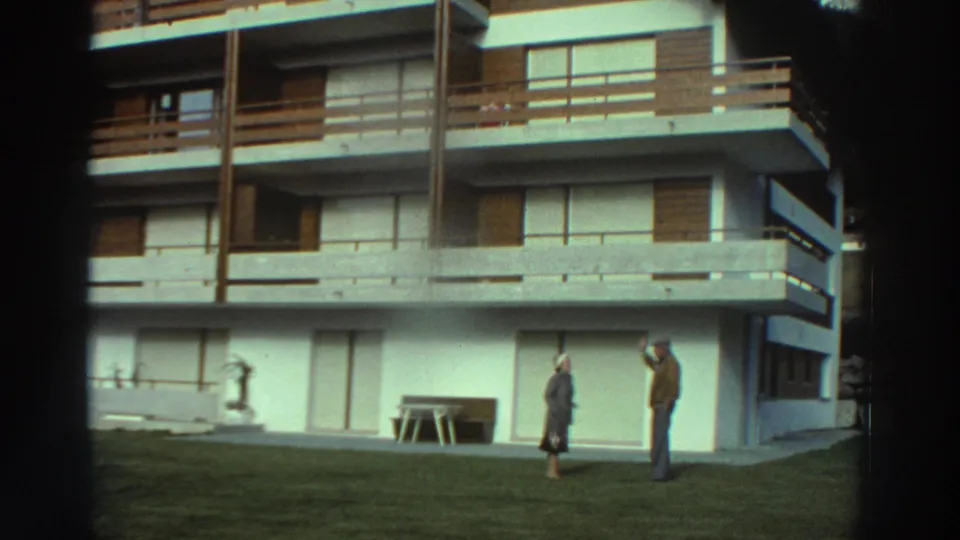 1976 Greece home-movie frame showing a modern apartment block with balconies, shutters, lawn, and two distant people outside.