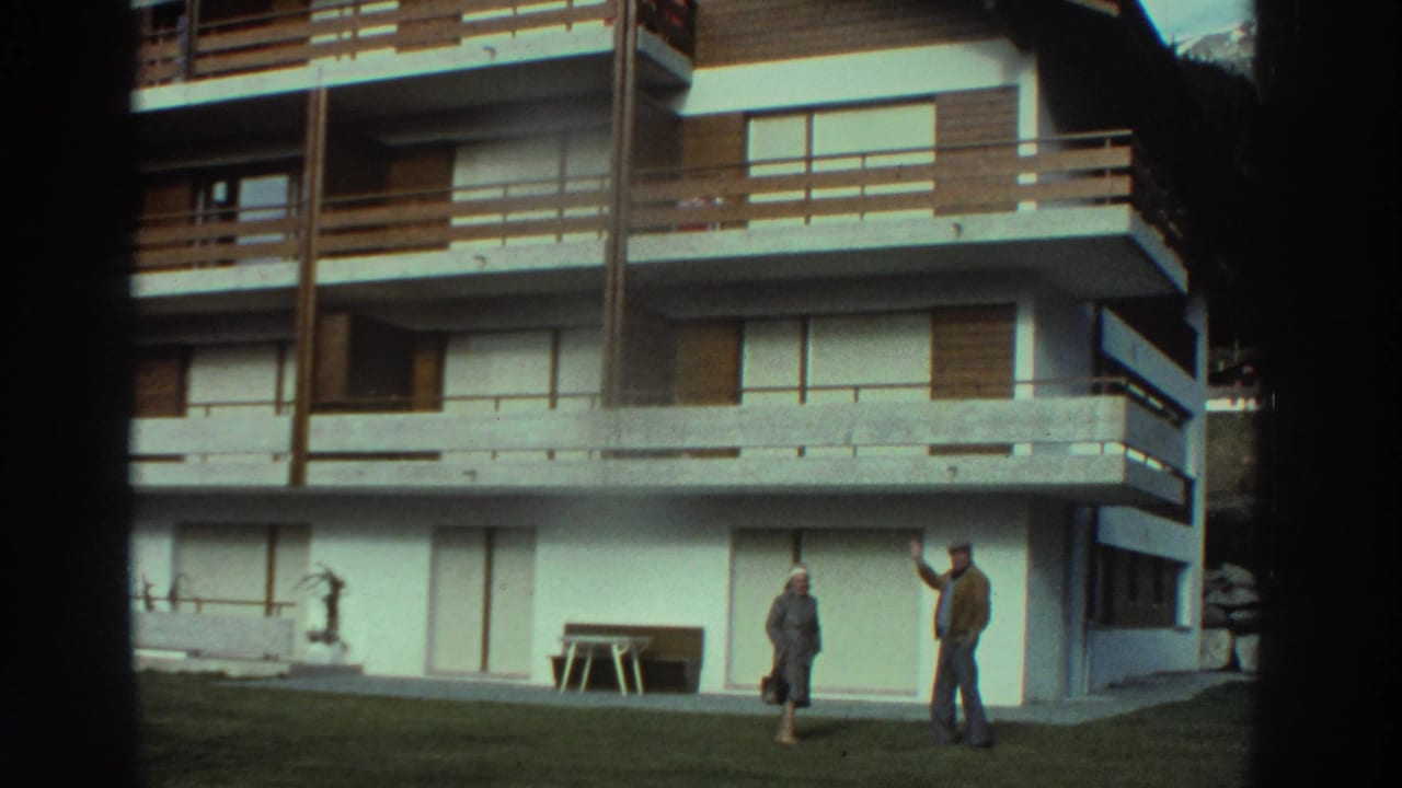 1976 Greece home-movie frame of two adults outside a white apartment or hotel building with timber balcony railings.