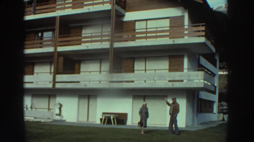 1976 Greece home-movie frame of two adults outside a white apartment or hotel building with timber balcony railings.