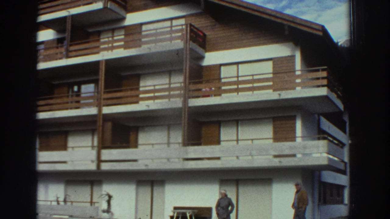1976 Greece frame showing the same white resort-style building, wooden balconies, and two adults partly separated across the lawn.