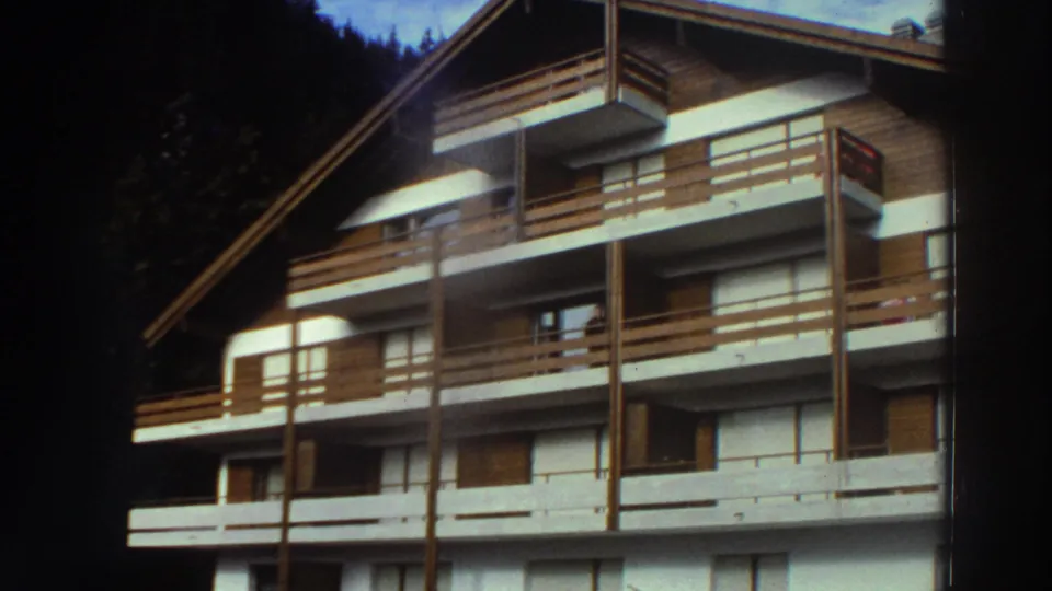 1976 Greece frame of a chalet-style apartment or hotel exterior with stacked wooden balconies and dark trees behind it.