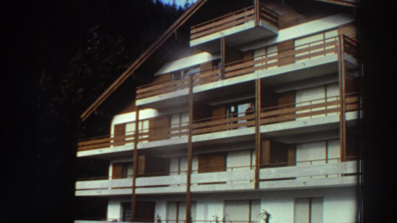 1976 Greece home-movie frame showing a white multi-story building with brown wood balcony rails and a steep roof near a wooded edge.