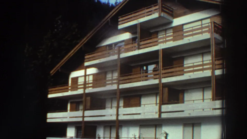 1976 Greece home-movie frame showing a white multi-story building with brown wood balcony rails and a steep roof near a wooded edge.