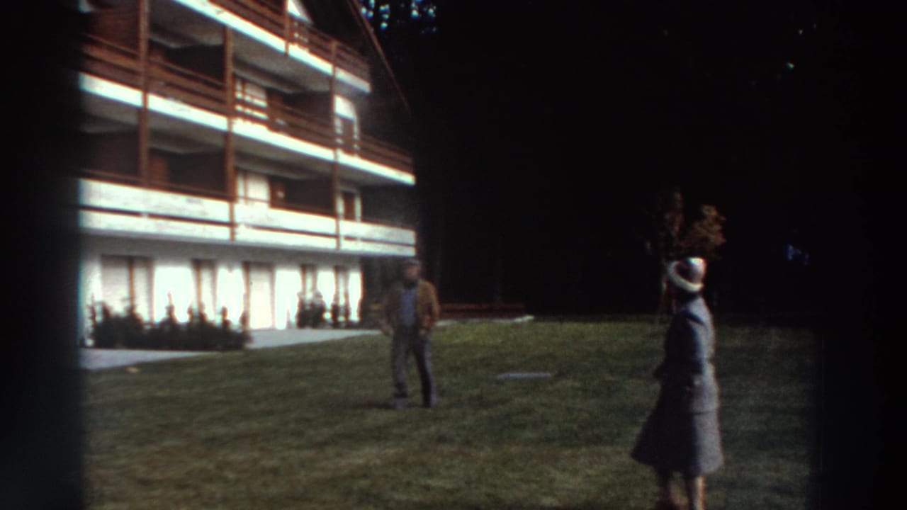 1976 Greece exterior frame with two adults on a lawn before a white building with timber balconies and curtained windows.