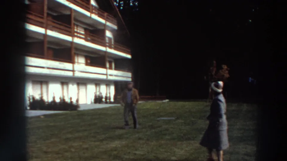 1976 Greece exterior frame with two adults on a lawn before a white building with timber balconies and curtained windows.