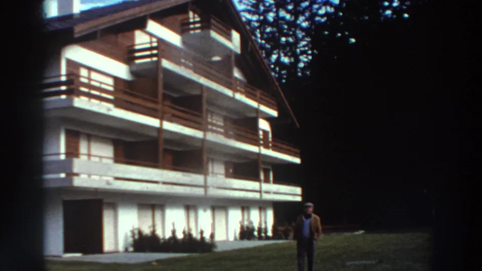 Home-movie frame from 1976 Greece showing a timber-balcony building, lawn, shrubs, and an adult standing near the facade.