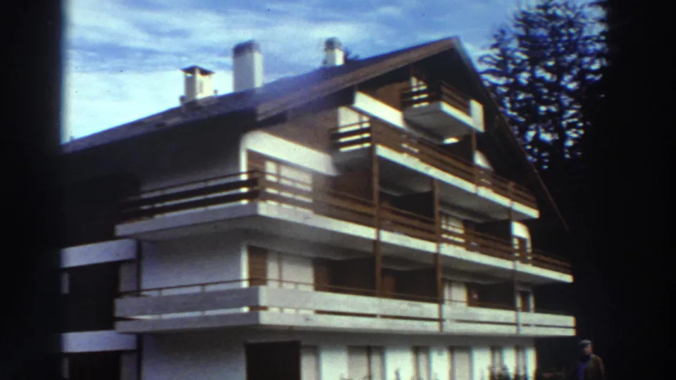 1976 Greece frame showing an angled exterior view of a white building with brown timber balconies, chimneys, and trees behind it.