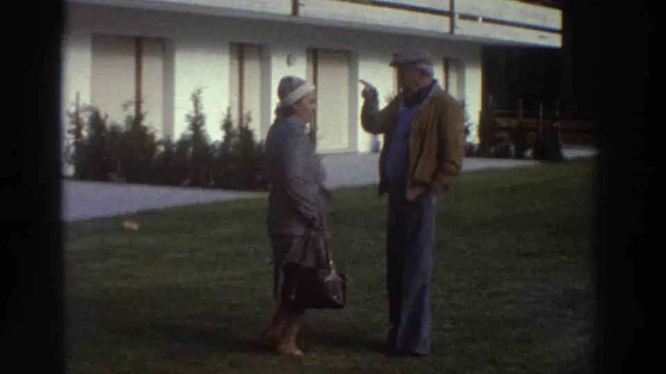 In front of the chalet, an elderly woman in a gray coat and hat faces an elderly man in a brown jacket and cap as he gestures.