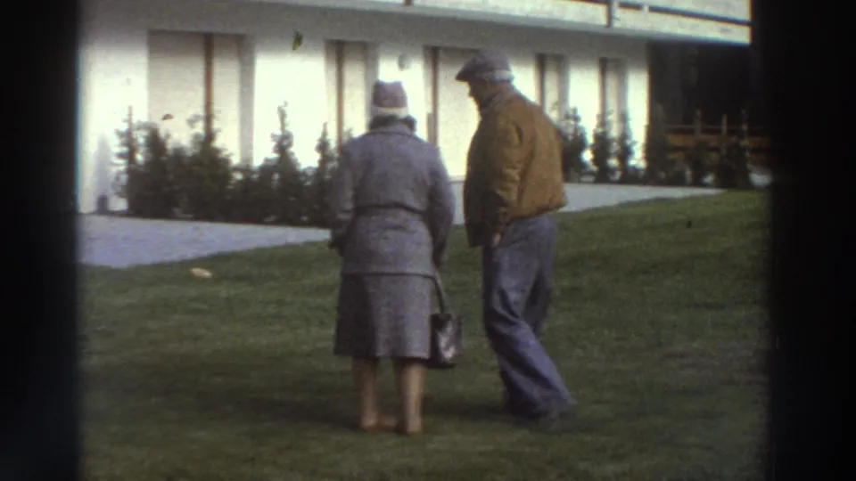 The elderly couple walks across the lawn beside the white chalet, seen from behind and in profile in soft 8mm color.