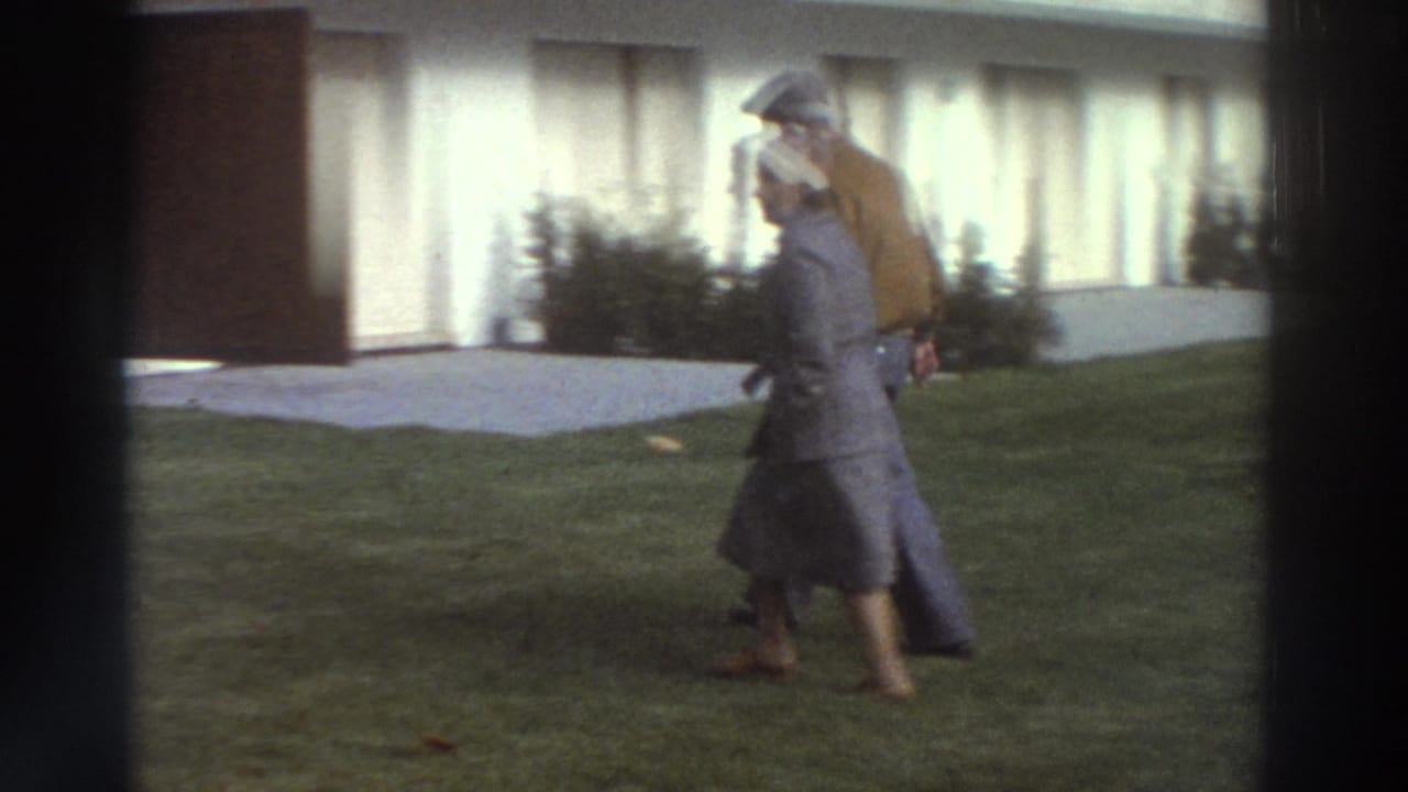 In 1976 Greece, two family members appear to walk across a lawn outside a white suburban house, filmed in soft 8mm color.