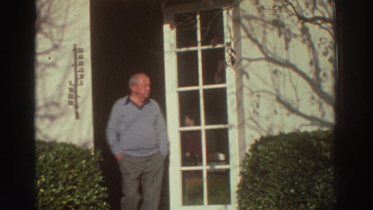 An older man stands in a doorway beside a grid-paned glass door and shrubs.
