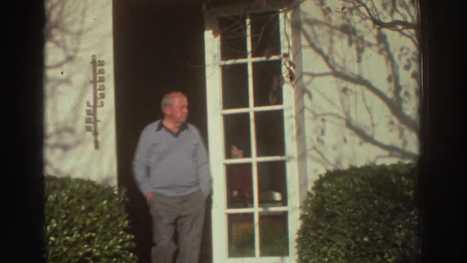 An older man in a light sweater stands at the doorway of a white house in Greece, with shrubs and branch shadows nearby.