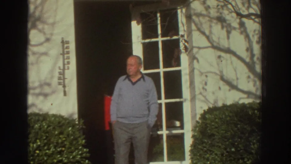 The older man remains near the house entrance, framed by the white wall, glass door, shrubs, and a partly visible red object.