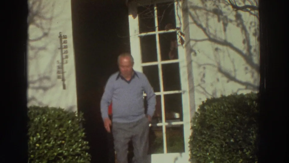 The older man stands near the grid-paned door and manicured shrub, looking down as bare branch shadows fall across the wall.