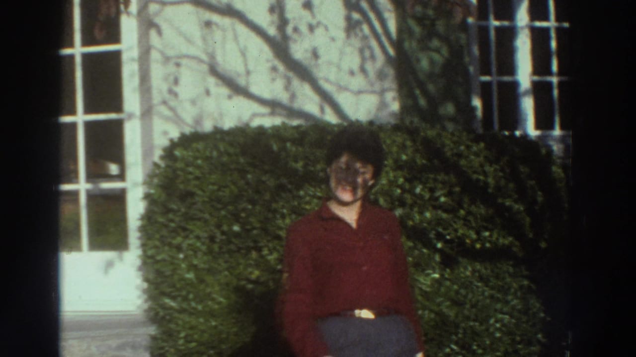 Adult in red stands near clipped hedges outside a white building in 1976 Greece home-movie footage.