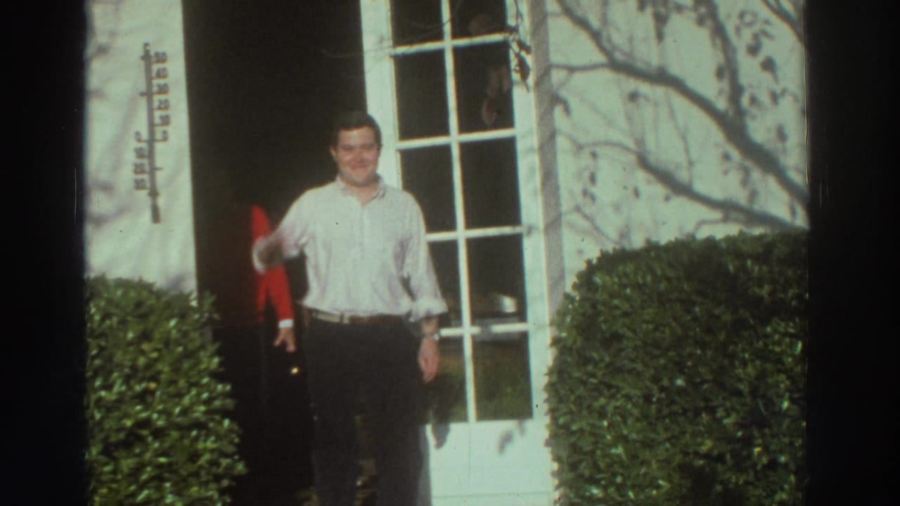 Man in a white shirt waves from the entrance of a white building in Greece, with hedges and windows behind him.