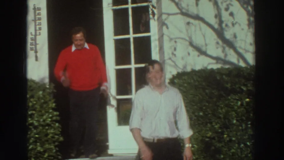 Two men stand outside the building entrance, one in a red sweater and one in a white shirt, in 1976 Greece footage.