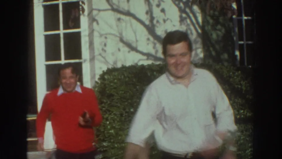 Two smiling men walk toward the camera outside a white building with clipped hedges in 1976 Greece home-movie footage.