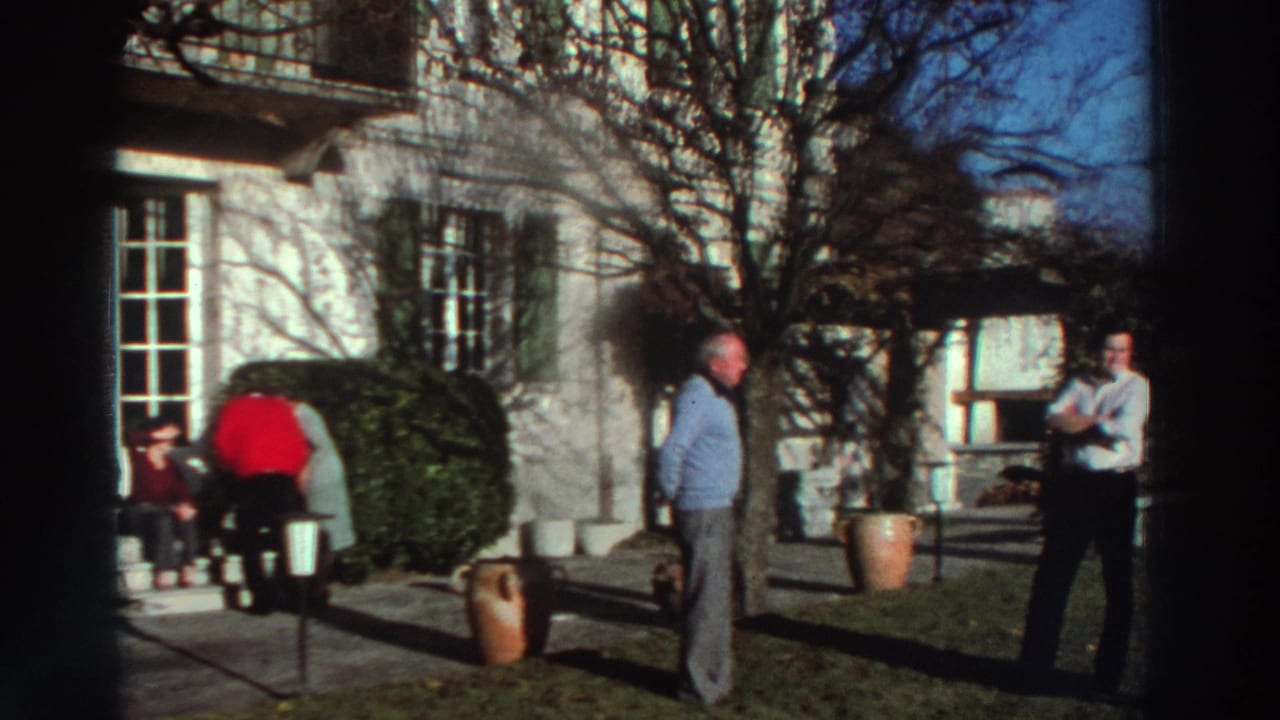 1976 Greece home-movie frame showing adults in a garden outside a pale stone house with green shutters, bare trees, and terracotta pots.