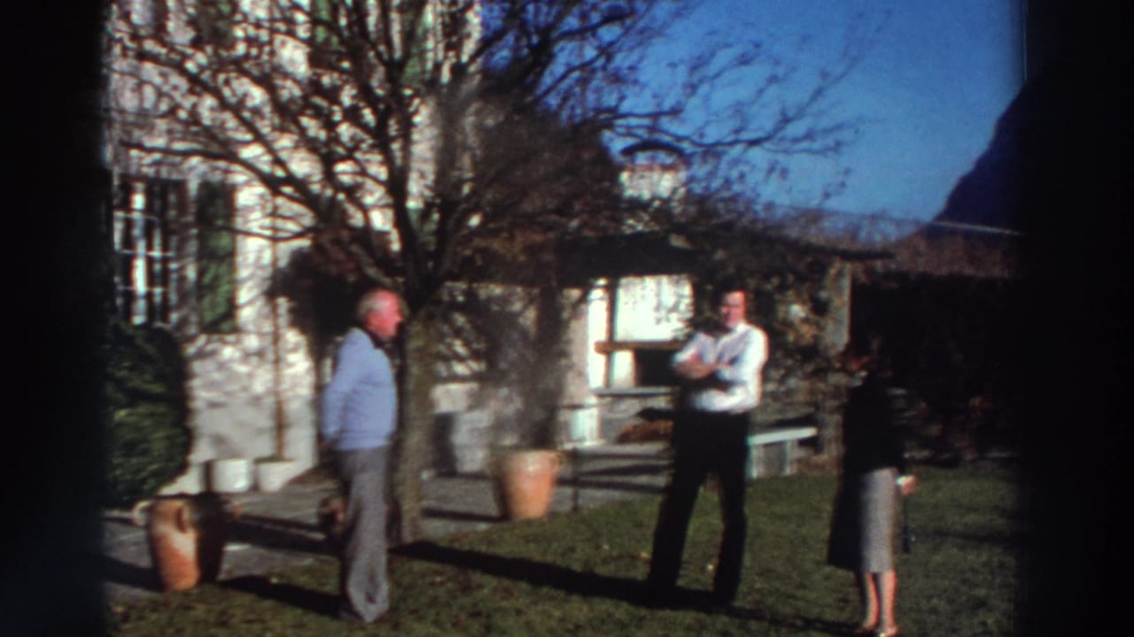Two men and a woman stand on a lawn near a shuttered house and garden pots.