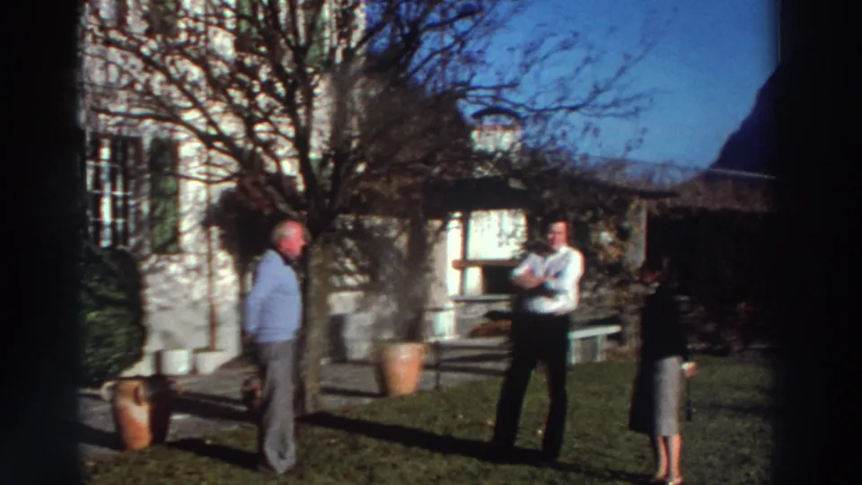 1976 Greece garden scene with two men and a woman near a pale house, terracotta pots, bare branches, and strong sunlight.