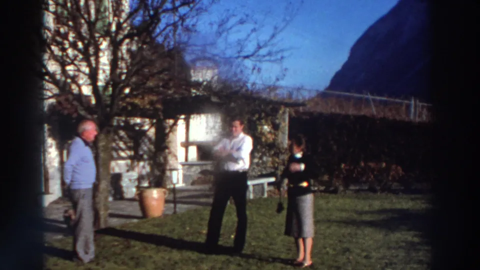 1976 Greece 8mm frame of adults in conversation on a sunlit lawn beside a shuttered stone house and garden pots.