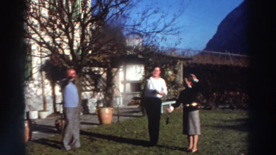 1976 Greece home-movie still showing three adults talking in a garden with a pale house, bare tree, pots, and distant hillside.