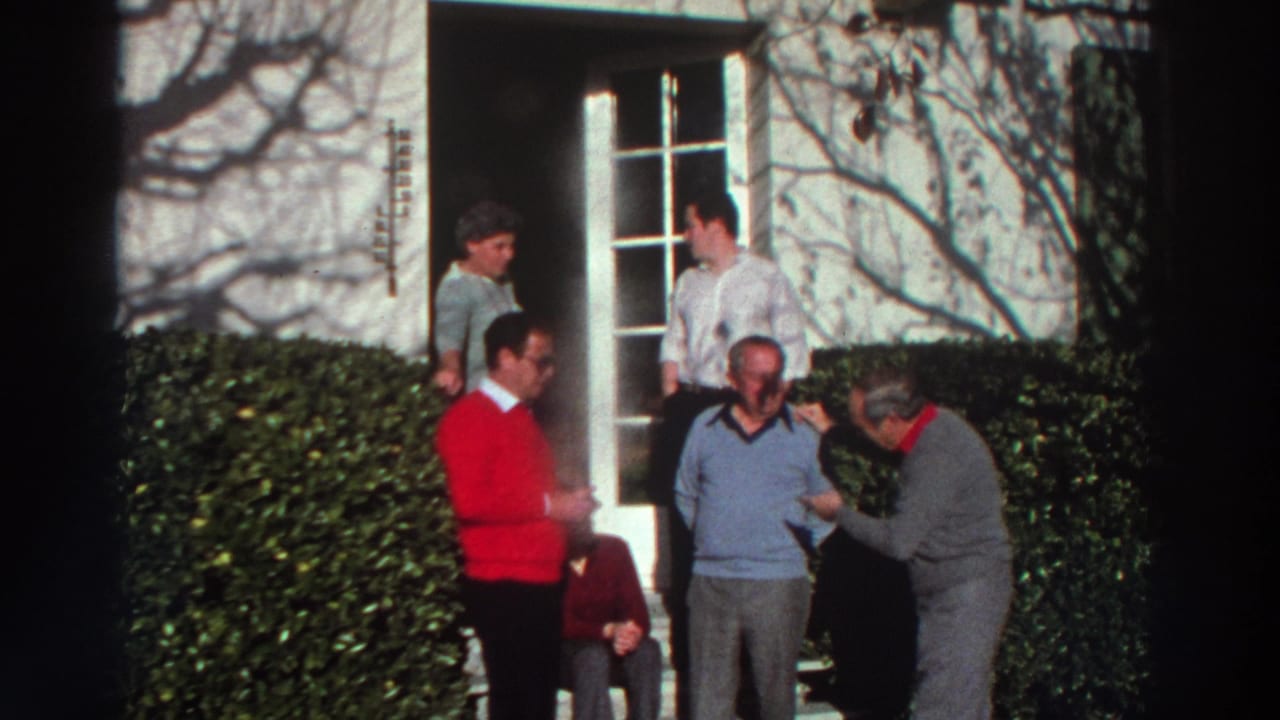 1976 Greece home-movie frame showing adults on front steps near a doorway, with hedges and branch shadows on a white facade.