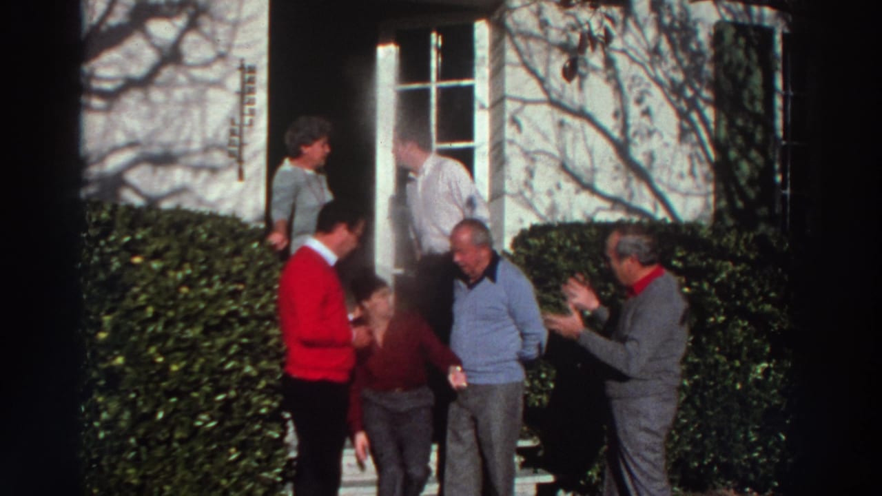 Group of adults converse on steps outside a house doorway with hedges in front.