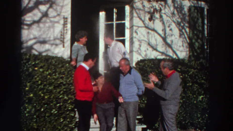 1976 Greece home-movie frame of adults conversing and shifting positions on exterior steps beside hedges and a white doorway.