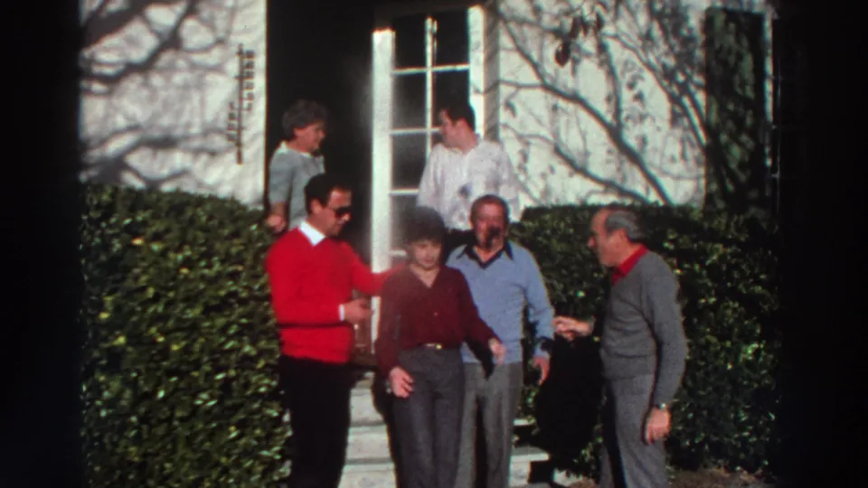 1976 Greece home-movie frame showing adults clustered near a doorway, including a man in a red sweater and a person in a maroon top.
