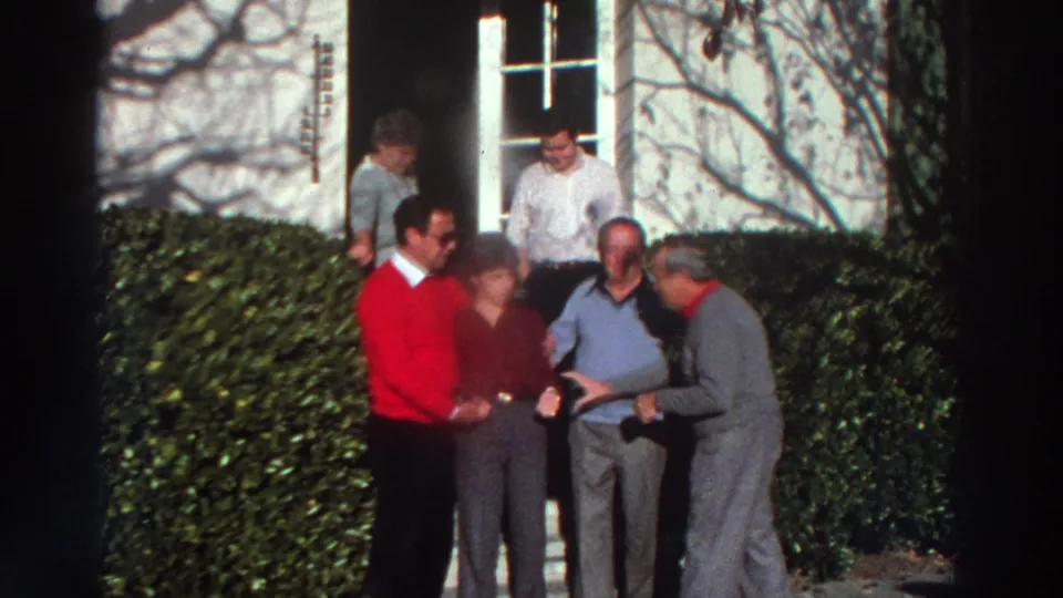 1976 Greece home-movie frame of adults gesturing and talking outside a house entrance, framed by hedges and shadows on the wall.