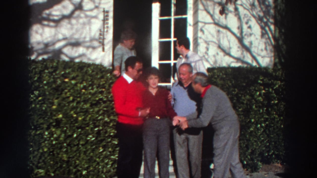 Adults gathered outside a white house, with one man in a red sweater embracing a woman.
