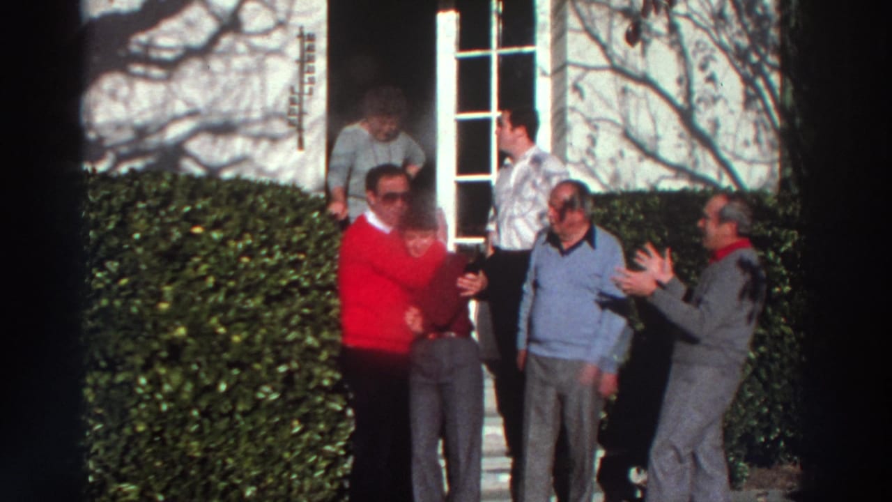 1976 Greece home-movie frame showing adults gathered near a doorway and hedges, with one man in a red sweater facing the group.