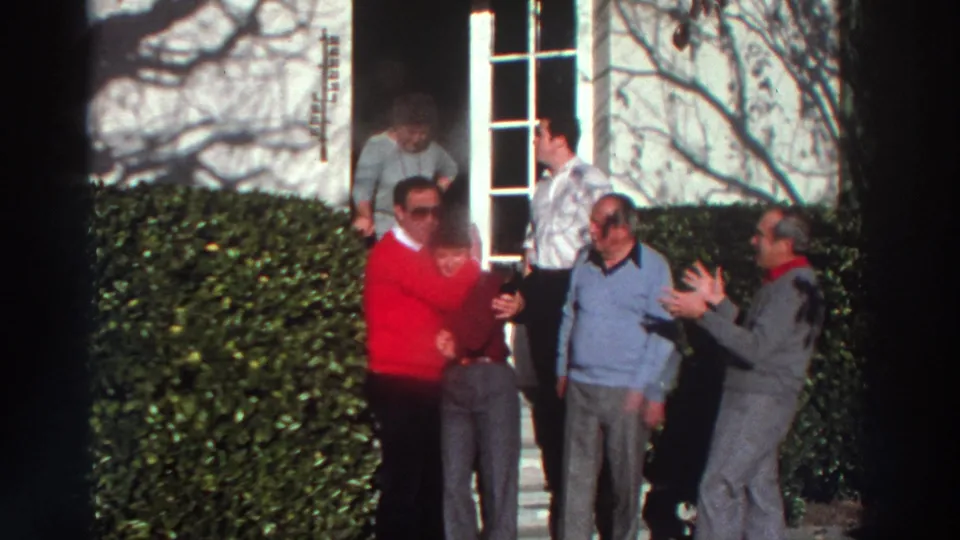 1976 Greece home-movie frame showing adults gathered near a doorway and hedges, with one man in a red sweater facing the group.