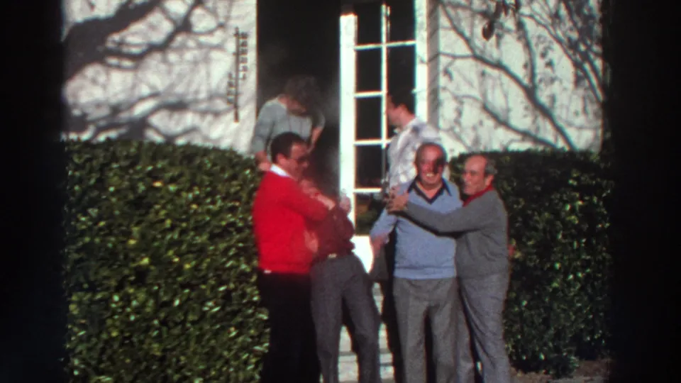 1976 Greece 8mm frame of adults laughing and hugging near a white house entrance, with hedges and strong sunlight visible.