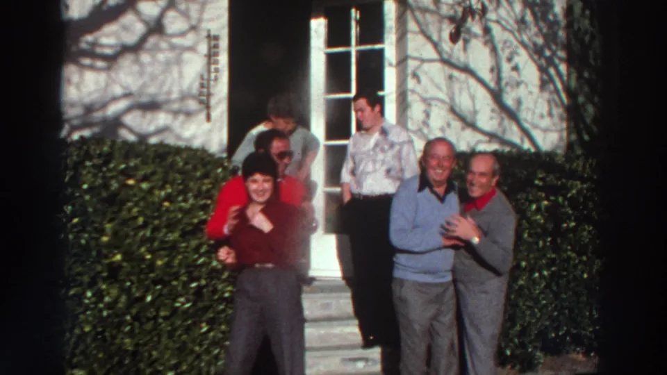 1976 Greece home-movie frame of adults smiling and standing together outside a white house, with one pair hugging in front.
