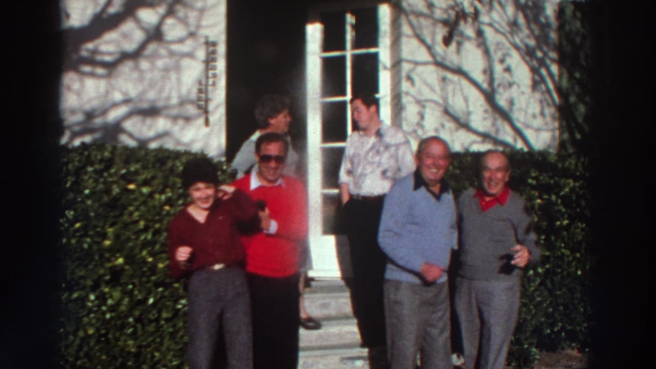 Adults in 1970s Greece stand together outside a doorway, smiling and posing on a sunlit porch with hedges nearby.