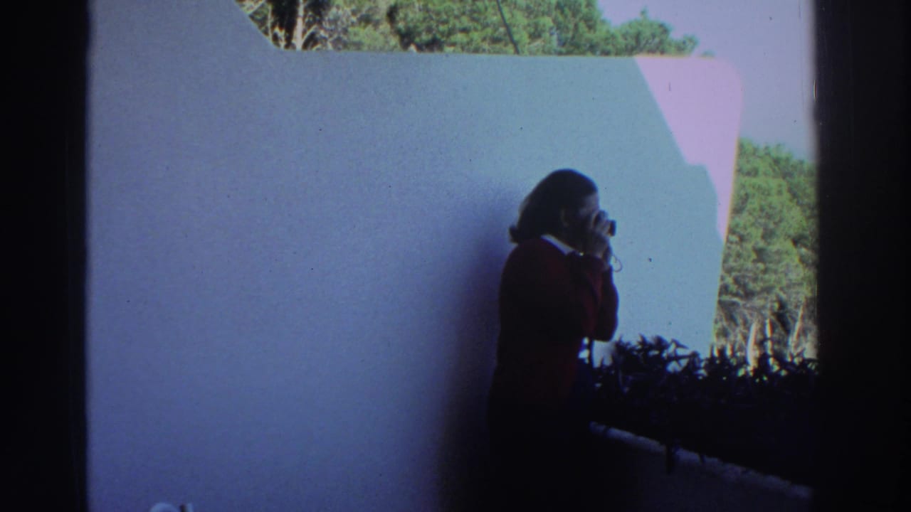 A woman in a red sweater appears in silhouette while using a camera near a white wall and shaded overlook in Greece.
