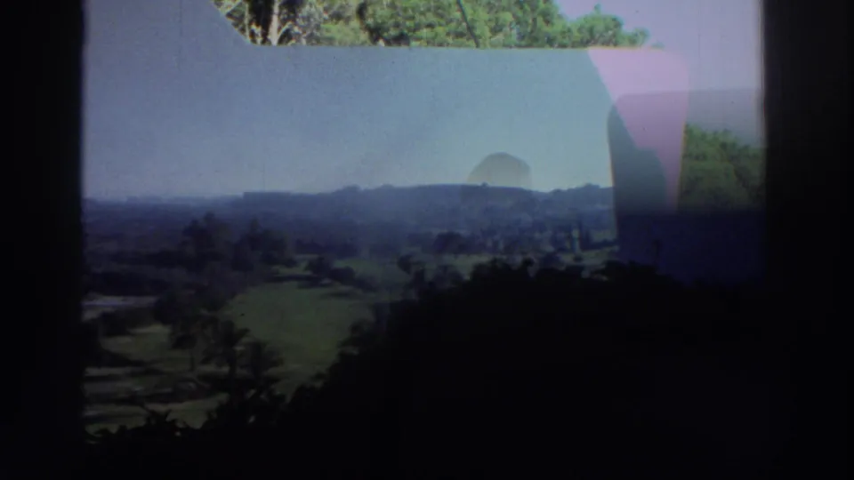 A shaded 8mm view looks past a white porch wall toward fields, trees, and low hills in Greece.