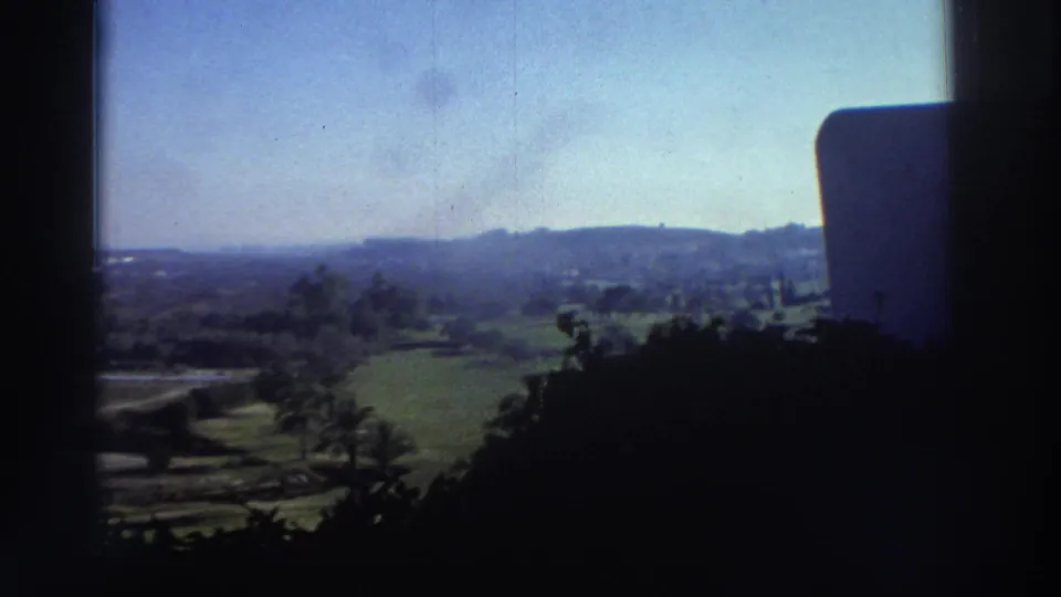 A soft 8mm landscape view shows fields, trees, and hills beyond a shaded porch or terrace in Greece.