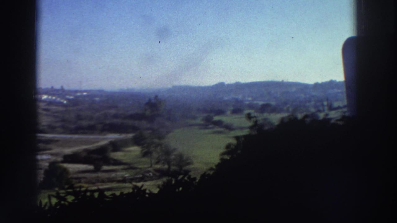 Landscape seen through dark window edges with hills, green fields, roads, and distant buildings.