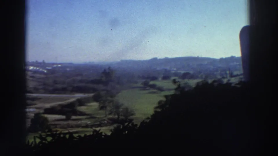 1976 8mm Greece home-movie frame looking through a window toward hazy hills, green fields, roads, and a distant built-up horizon.