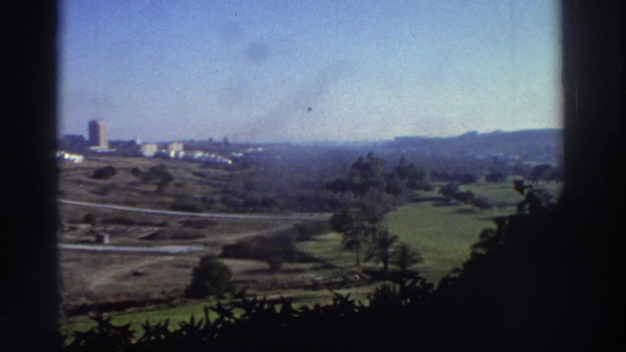 1976 Greece home-movie view across green fields and roads toward distant buildings and low hills, partly framed by dark window edges.