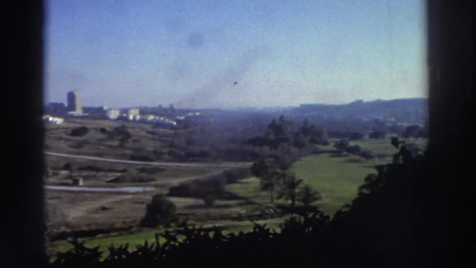 1976 Greece home-movie view across green fields and roads toward distant buildings and low hills, partly framed by dark window edges.