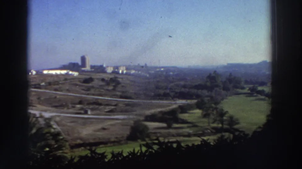 1976 8mm Greece frame showing a distant built-up area, roads, trees, and green fields through dark window or balcony framing.