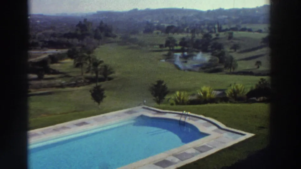 1976 Greece home-movie frame with a bright blue swimming pool, manicured lawn, palms, fields, water feature, and hazy hills beyond.