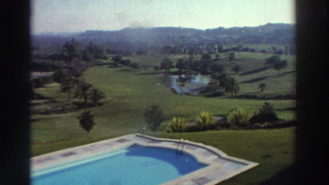 1976 Greece 8mm frame showing a pool in the foreground, lawns, trees, a small pond, and distant hills through a window frame.