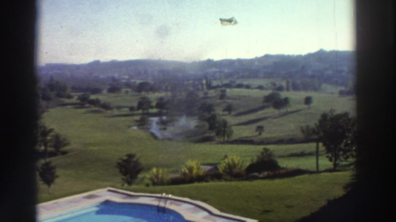 1976 Greece home-movie view with the pool edge below, landscaped grass, scattered trees, a small pond, and hazy hills in the distance.