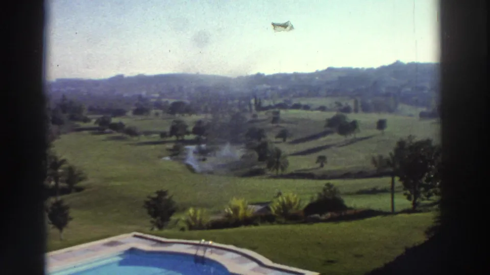 1976 Greece home-movie view with the pool edge below, landscaped grass, scattered trees, a small pond, and hazy hills in the distance.